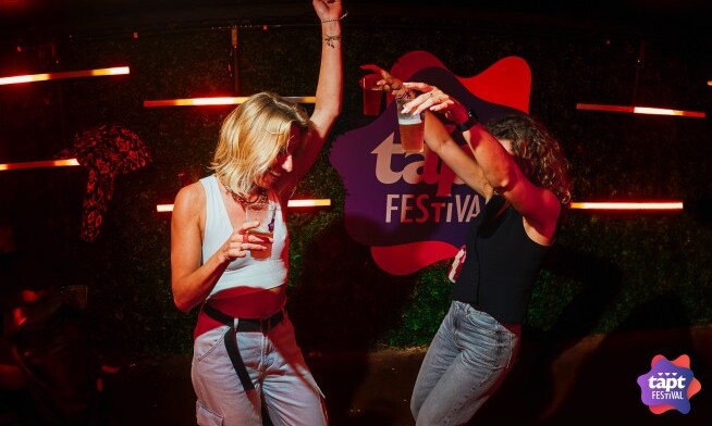 Twee mensen dansen met drankjes in hun hand voor een TAPT Festival bord.