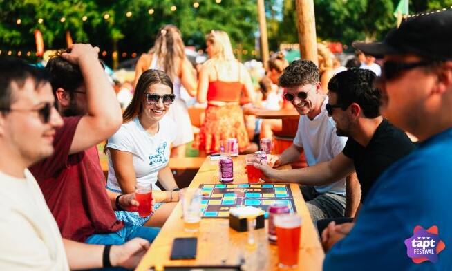 Groep mensen zit lachend aan een picknicktafel met drankjes en een spel bij TAPT Festival.