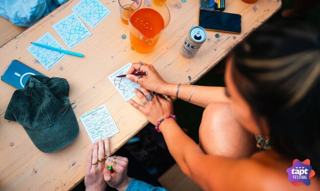 Personen spelen een kaartspel aan een tafel tijdens TAPT festival met drankjes en pet.