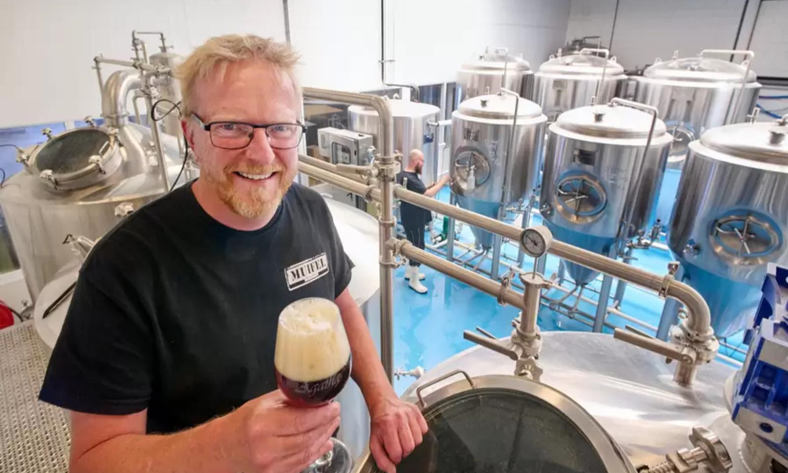 Brouwer Martin met glas bier in fabriek met roestvrijstalen tanks.