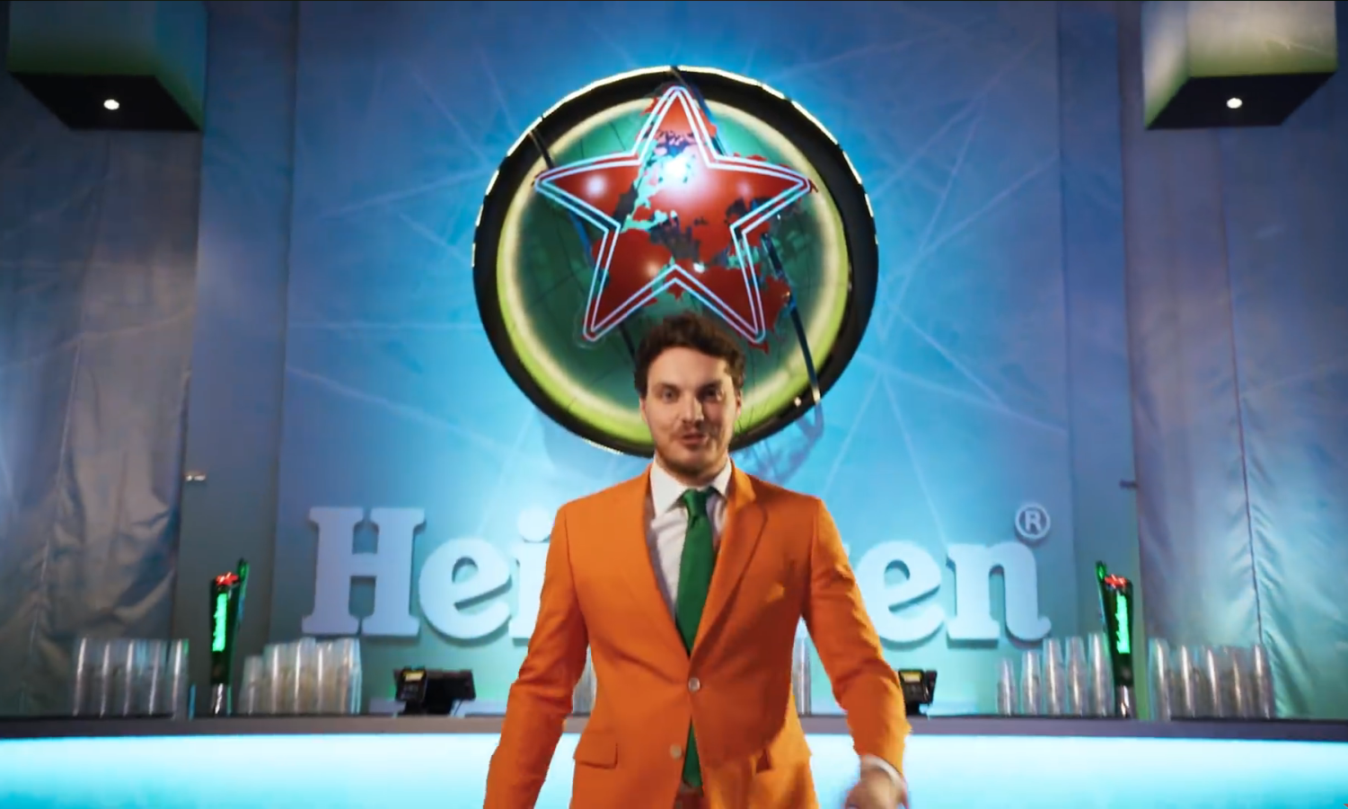 Een man in een oranje pak met groene stropdas staat voor het Heineken-logo.