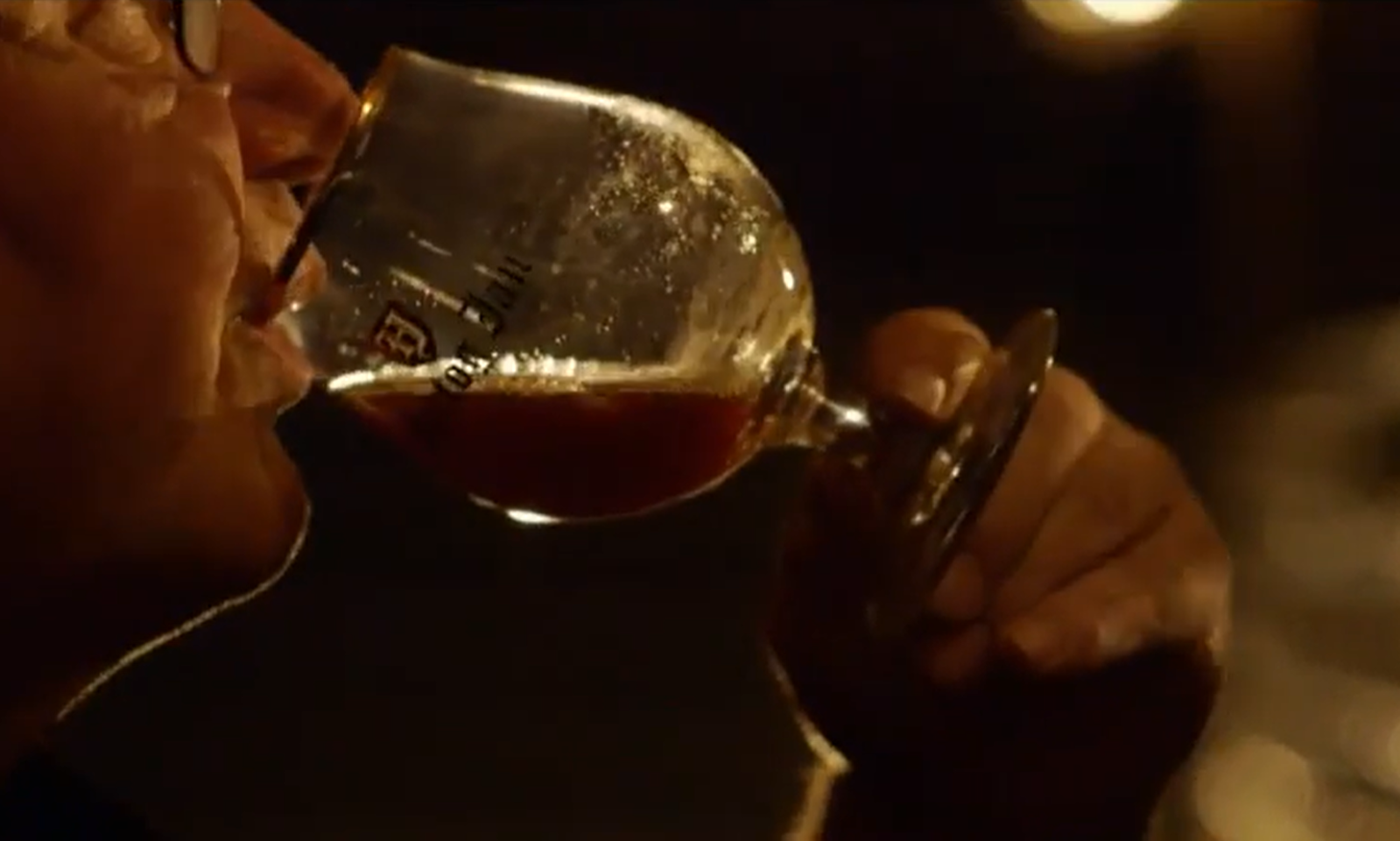 Man drinkt donker bier uit een glas met logo in een sfeervol verlichte ruimte.