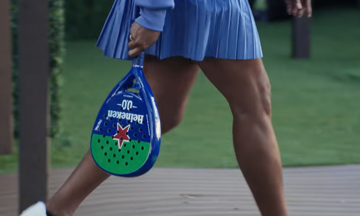 Persoon in blauwe rok met Heineken padelracket op grasveld.